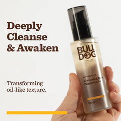 A hand holds a bottle of Anytime Daily Cleansing Face Concentrate. Text reads “Deeply Cleanse & Awaken” and “Transforming oil-like texture.” Made with Natural Origin Ingredients, the clear bottle features a black pump and gold label.